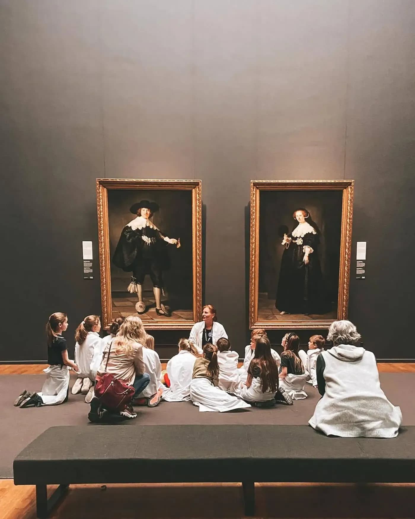 Group of schoolchildren listening to a guide in front of two classical portraits in a museum